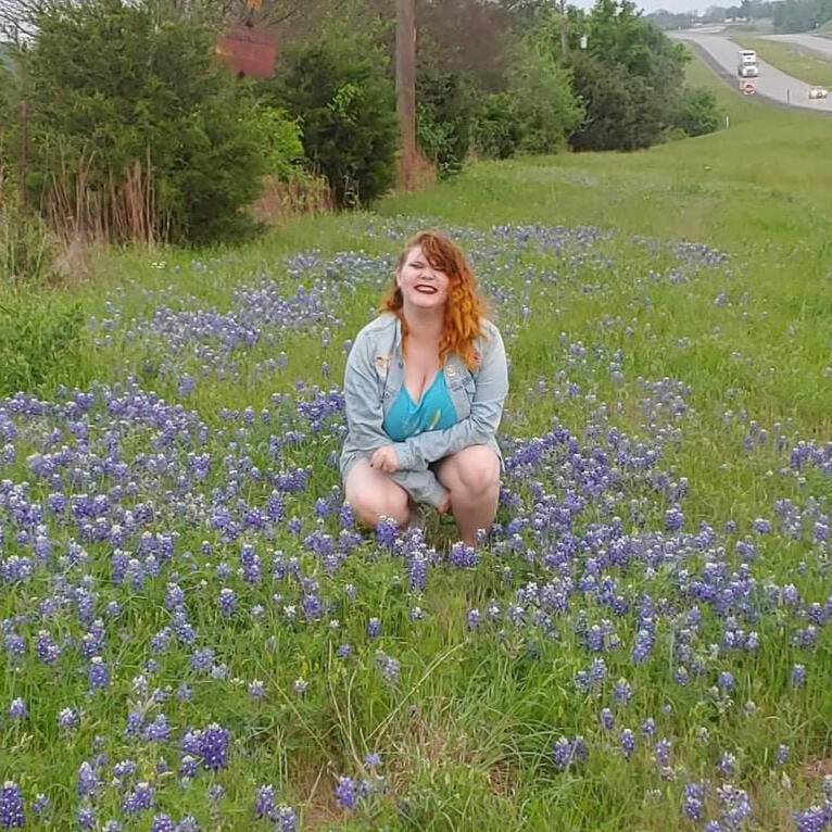 Photo of a redheaded nonbinary person (Mik) kneeling in a field of bluebonnets. They are smiling and have dark lipstick on.